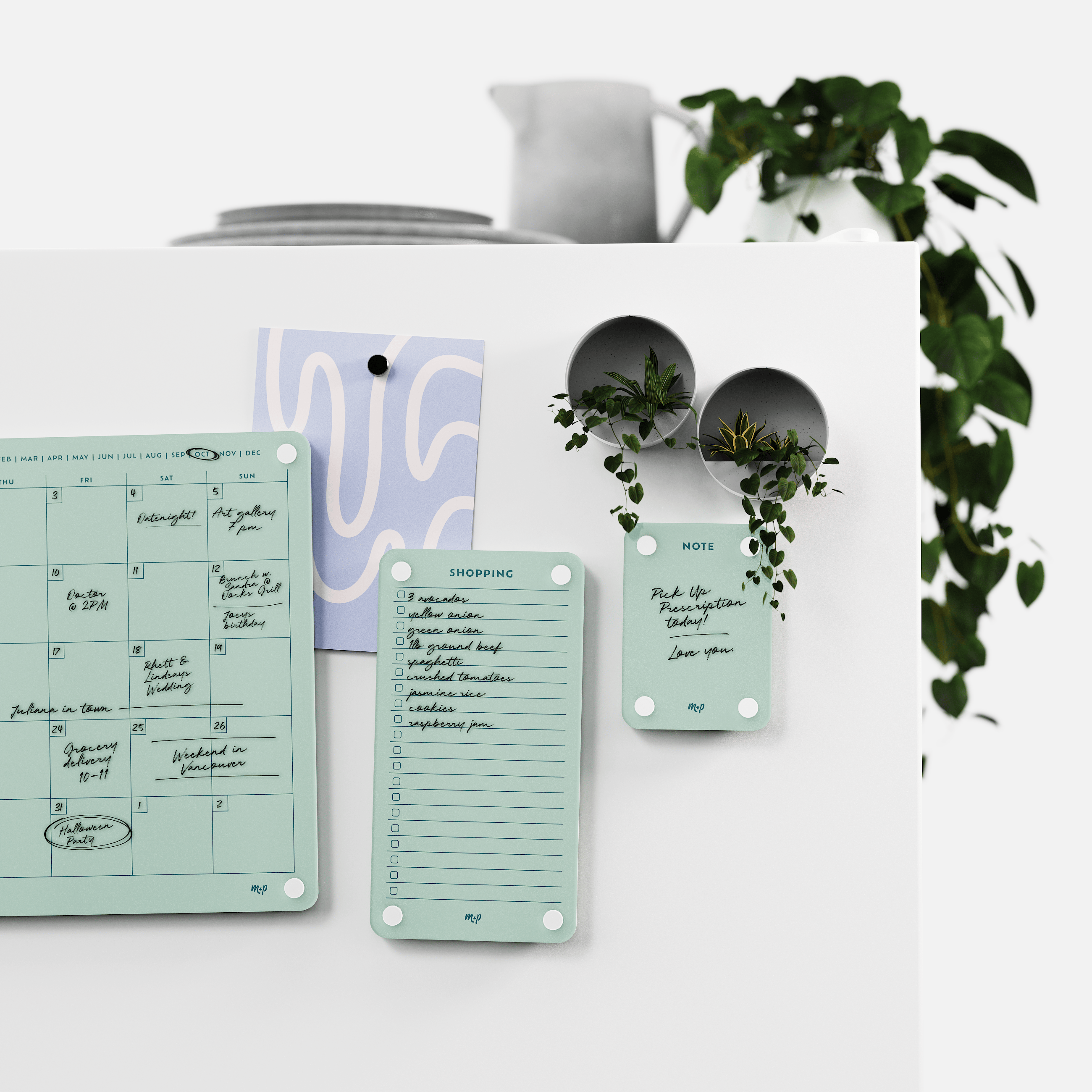 Seafoam green monthly calendar, shopping list and note board on a white kitchen wall with magnetic planters